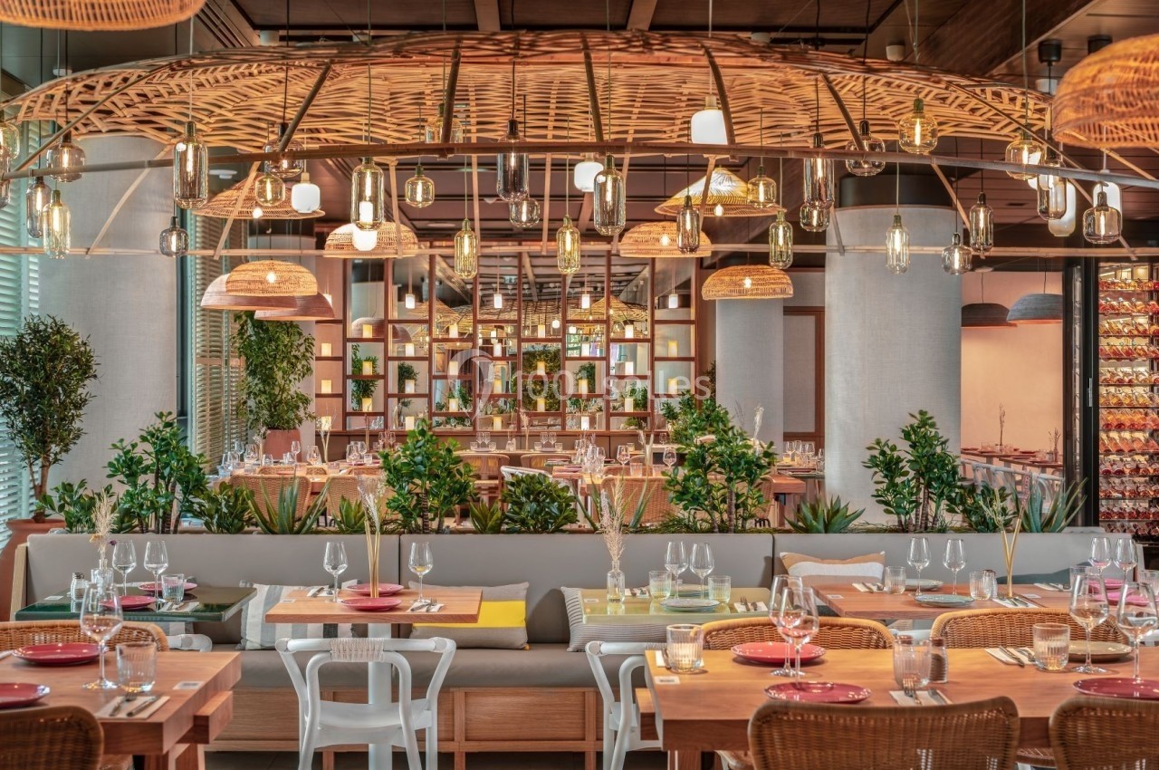 Salle de restaurant moderne avec tables dressées, chaises variées, plantes décoratives et éclairage suspendu en osier.
