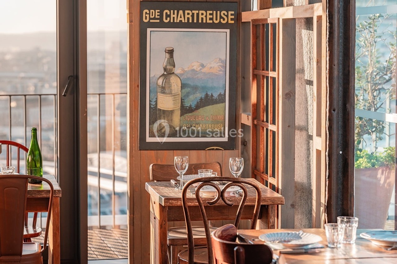 Salle de restaurant lumineuse avec des tables en bois, une affiche de la Chartreuse et une vue sur l'extérieur.