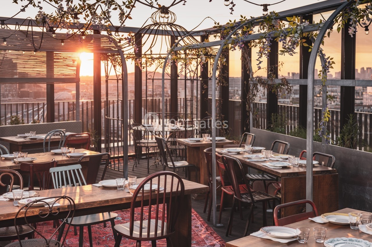 Terrasse de restaurant en rooftop au coucher du soleil, avec tables dressées, chaises variées et décoration végétale.