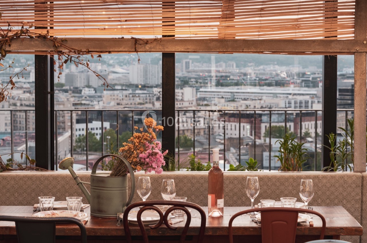 Table dressée avec des verres et une carafe, vue sur une ville à travers de grandes fenêtres et des stores en bois.
