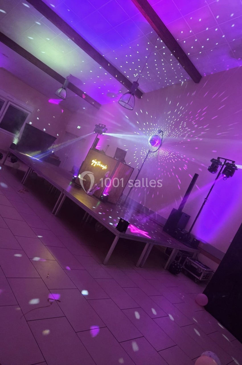 Salle de réception éclairée par des lumières colorées avec une scène, une boule disco et des équipements de sonorisation.