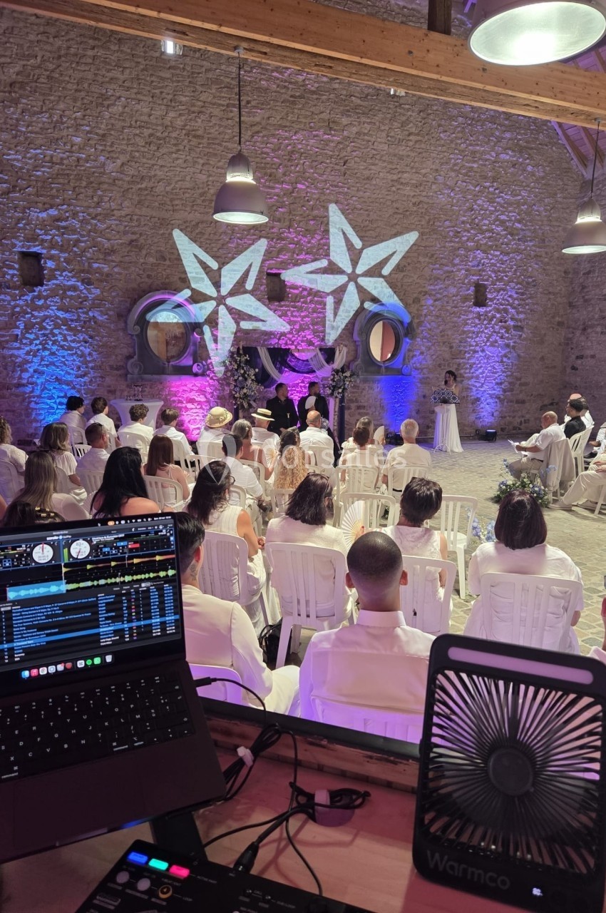 Cérémonie dans une salle en pierre avec invités assis, décor lumineux étoilé et matériel de DJ au premier plan.