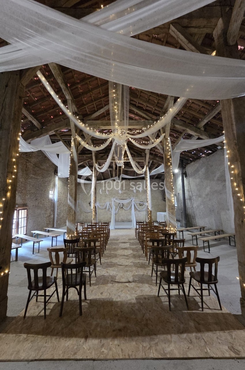 Salle de réception rustique avec poutres apparentes, guirlandes lumineuses et décoration en tissu blanc suspendu.