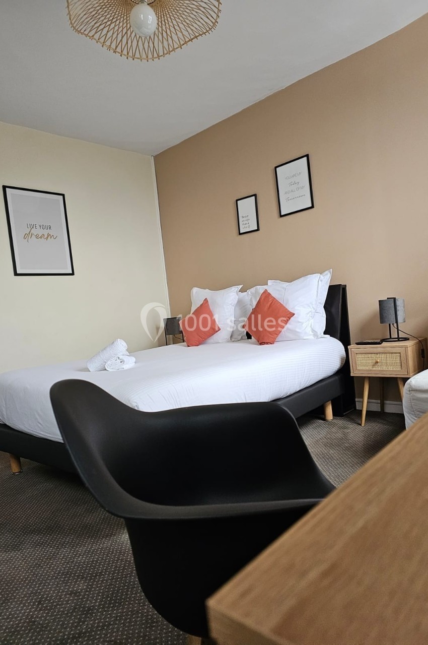 Chambre moderne avec lit double, coussins orange, murs beige et décor minimaliste, vue sur un bureau et un fauteuil noir.