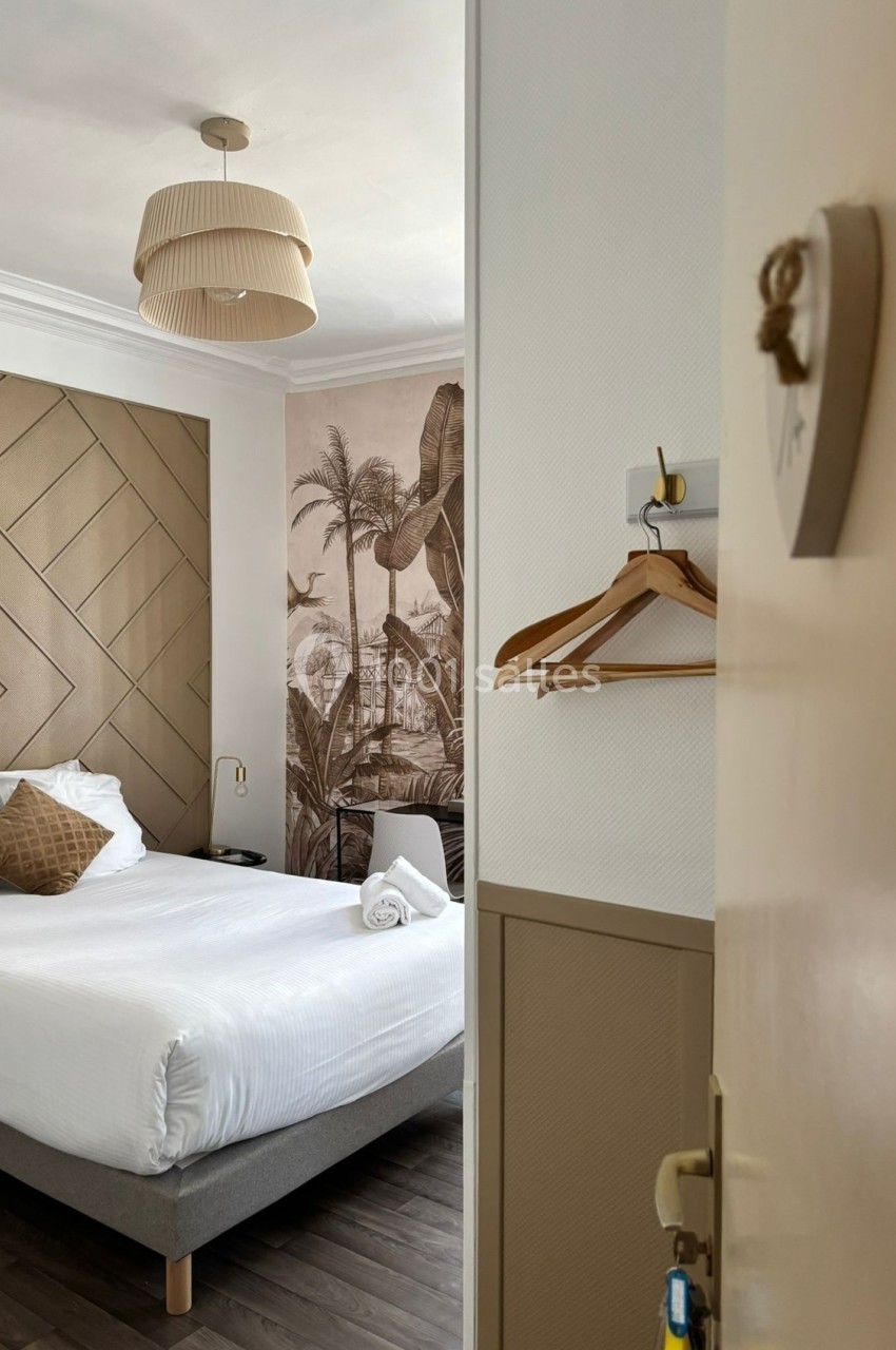 Chambre d'hôtel lumineuse avec lit double, linge blanc, décoration murale tropicale et cintres accrochés au mur.