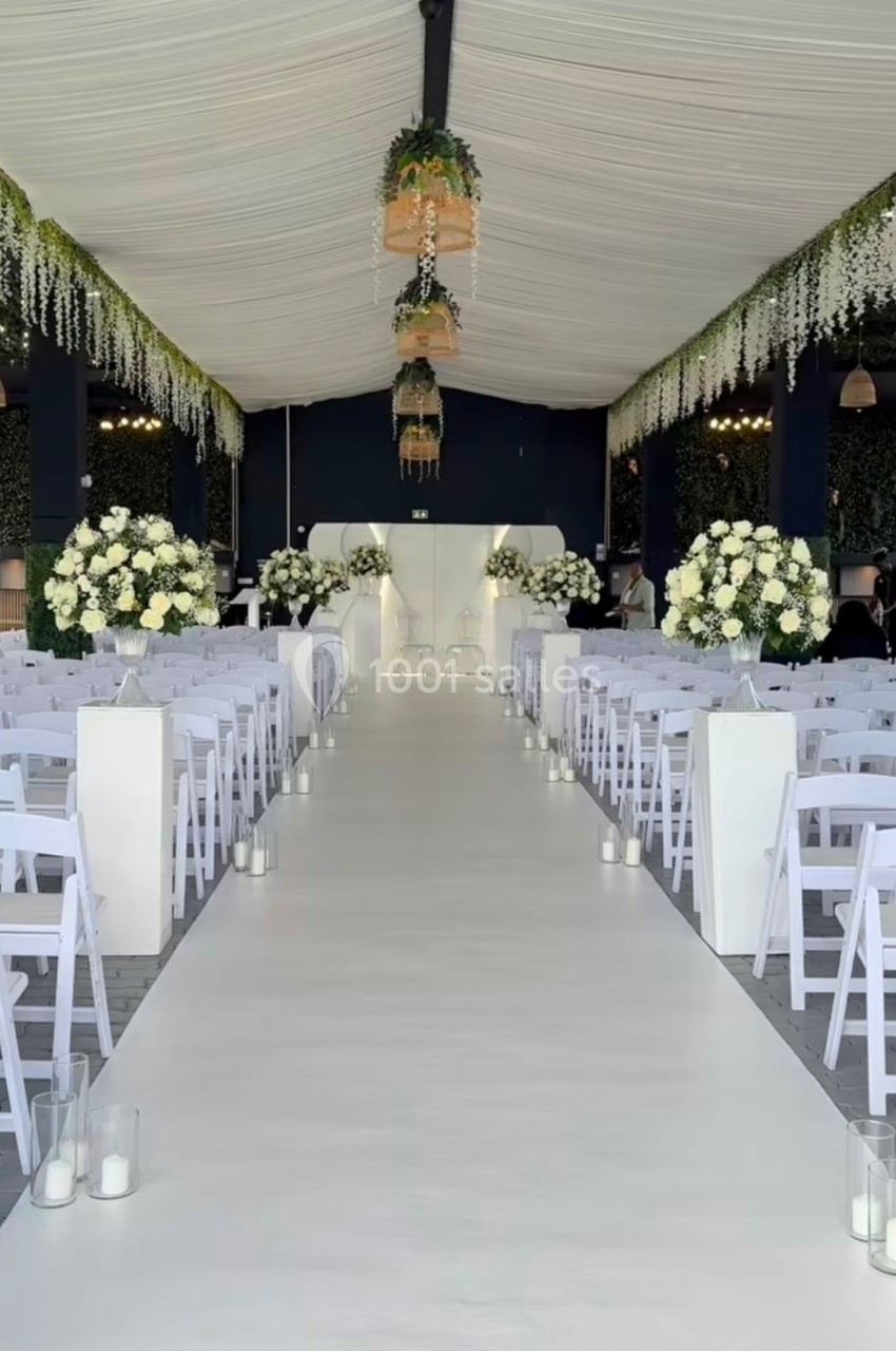 Allée centrale décorée de fleurs blanches et de guirlandes suspendues dans une salle de réception de mariage.