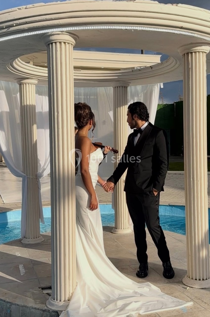Un couple en tenue de mariage se tient la main sous une structure circulaire avec des colonnes, près d'un bassin.