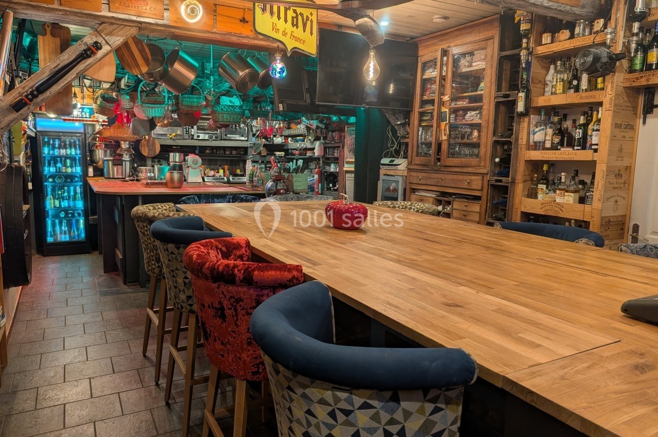 Grande table en bois entourée de chaises dans une cuisine ouverte avec étagères, ustensiles et réfrigérateur visible.