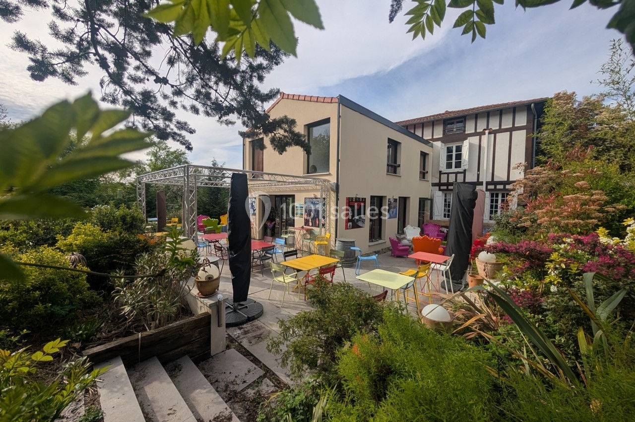 Terrasse d'un café avec tables colorées, entourée de végétation et jouxtant un bâtiment à colombages.