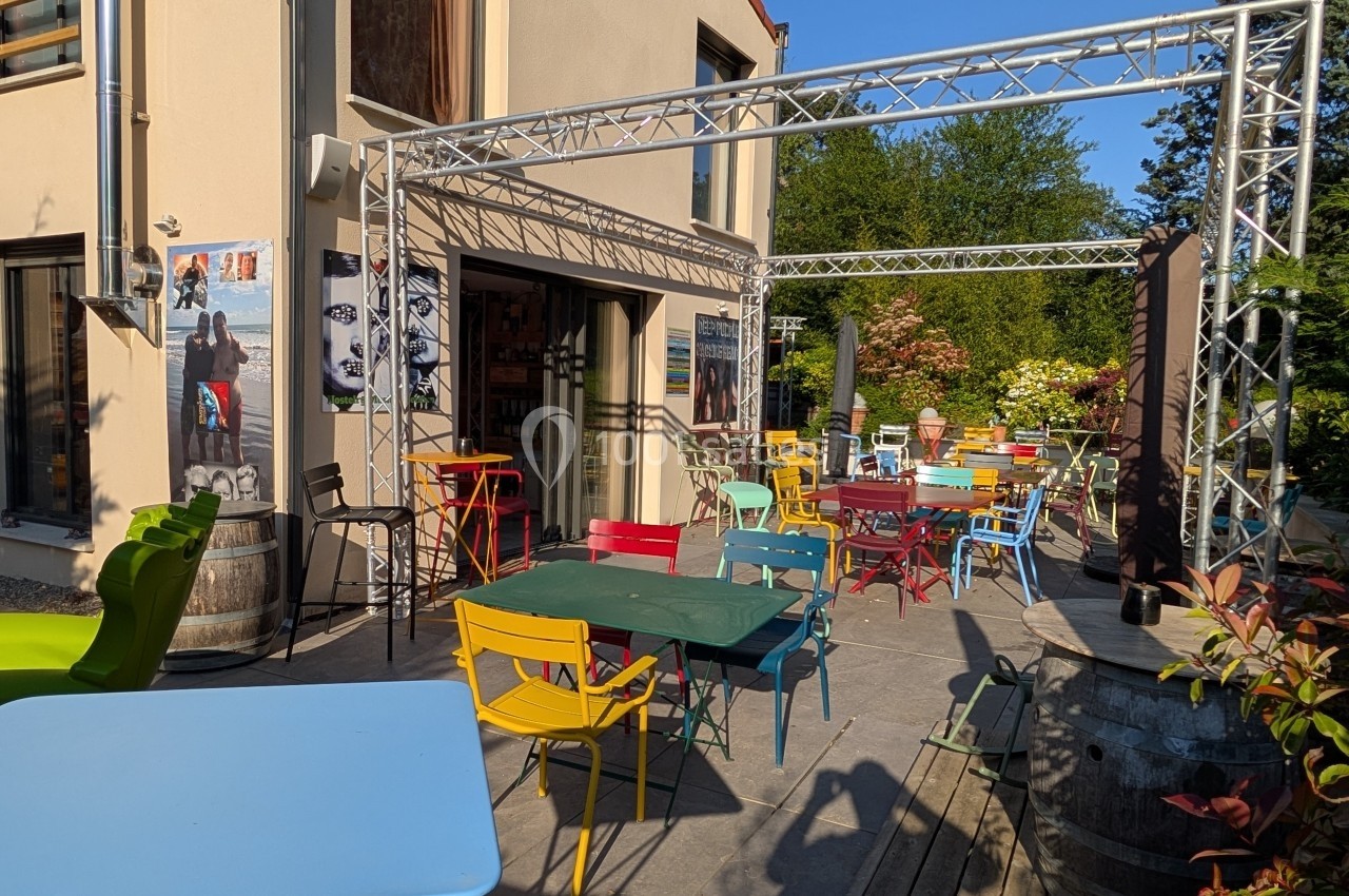 Terrasse ensoleillée avec tables et chaises colorées devant un bâtiment beige, entourée de verdure.