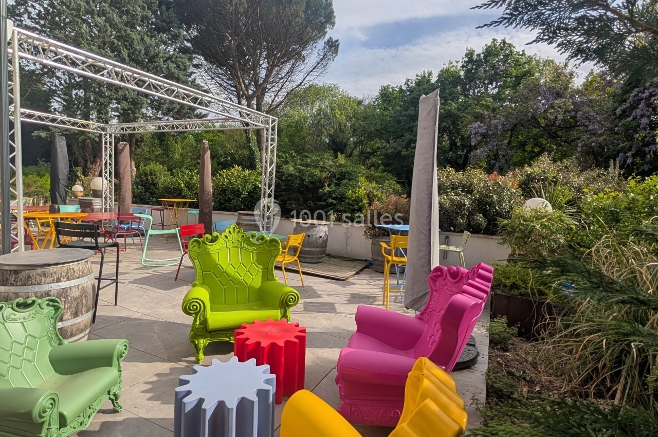 Terrasse extérieure avec fauteuils colorés, tables et végétation environnante sous un ciel partiellement nuageux.