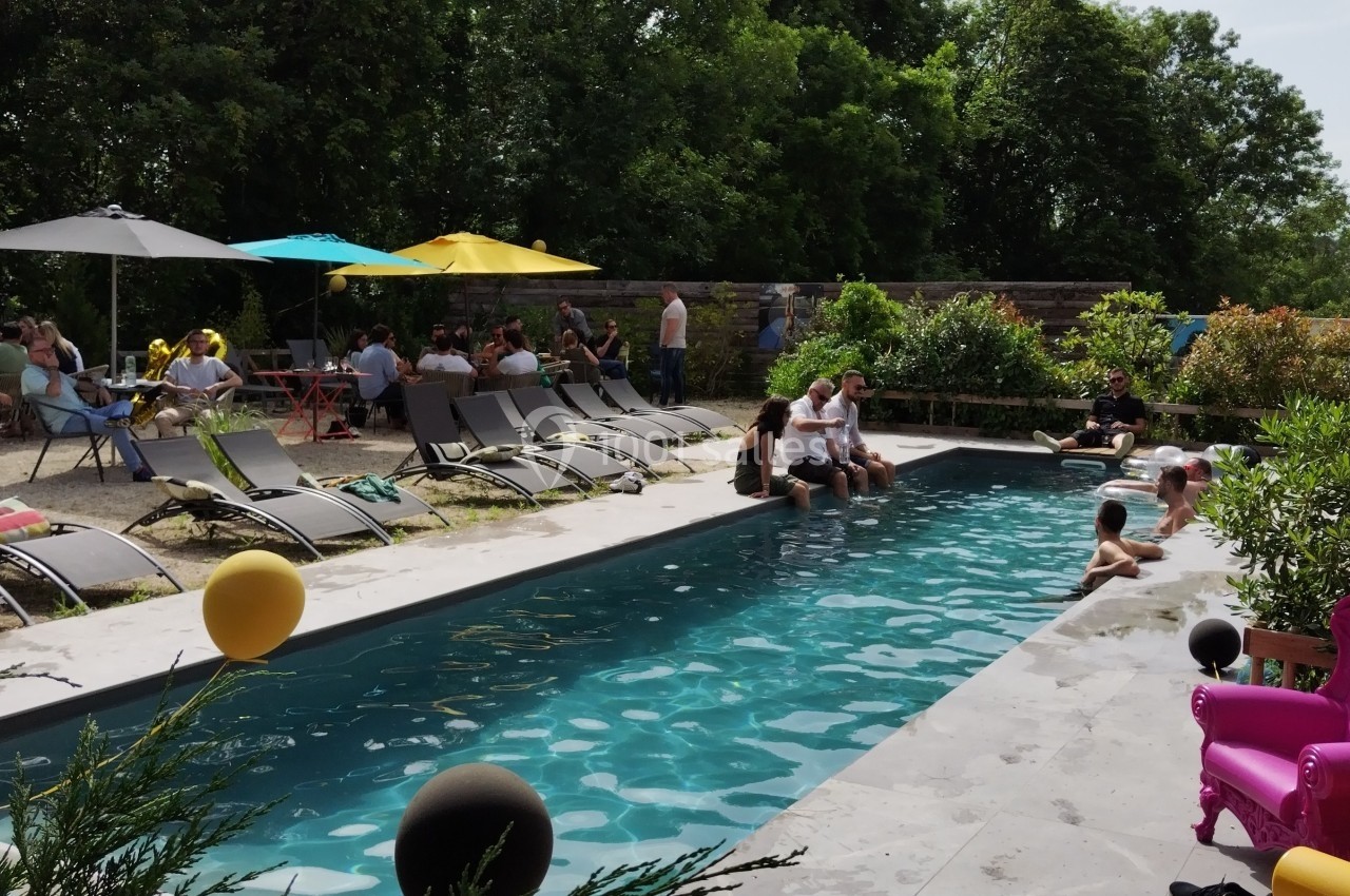 Personnes profitant d'une piscine extérieure entourée de chaises longues, parasols et végétation en arrière-plan.