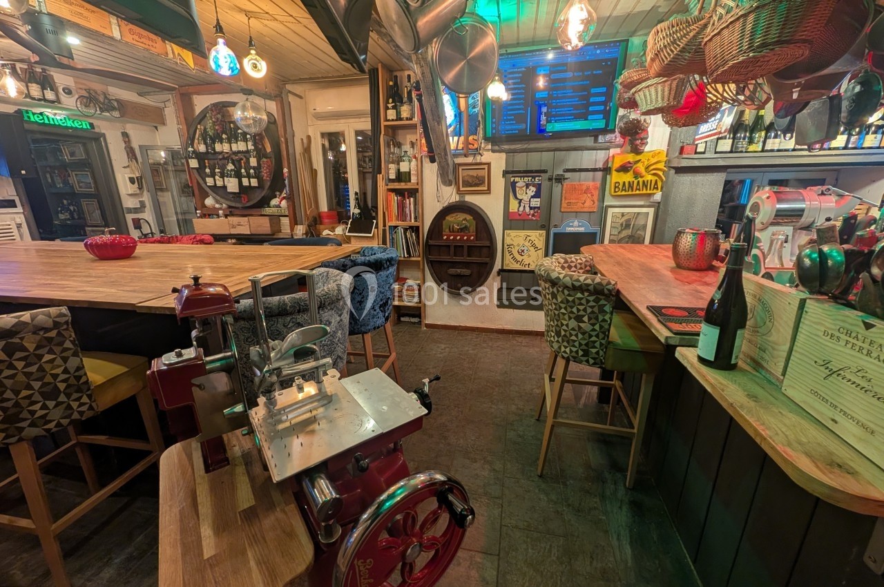 Intérieur d'un bar chaleureux avec des tables en bois, des affiches rétro, et une machine à trancher au premier plan.
