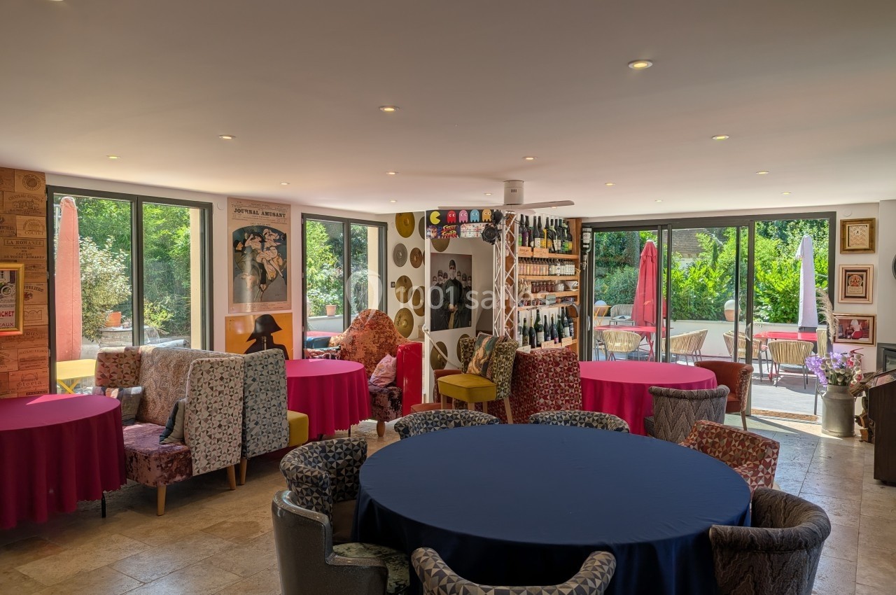 Salle lumineuse avec des tables rondes colorées, des fauteuils variés et des baies vitrées donnant sur un jardin.