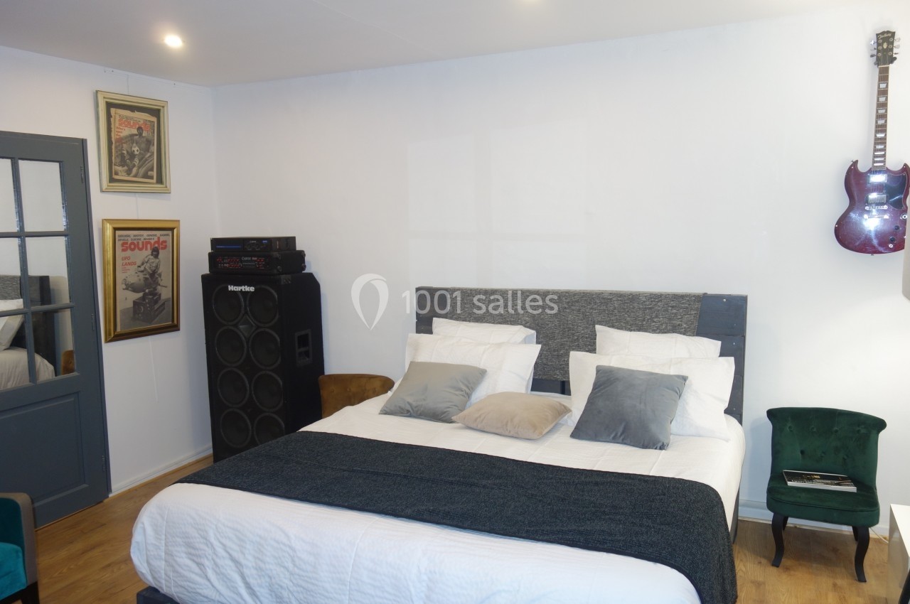 Chambre avec lit double, coussins décoratifs, guitare accrochée au mur et amplificateur à côté d'affiches encadrées.