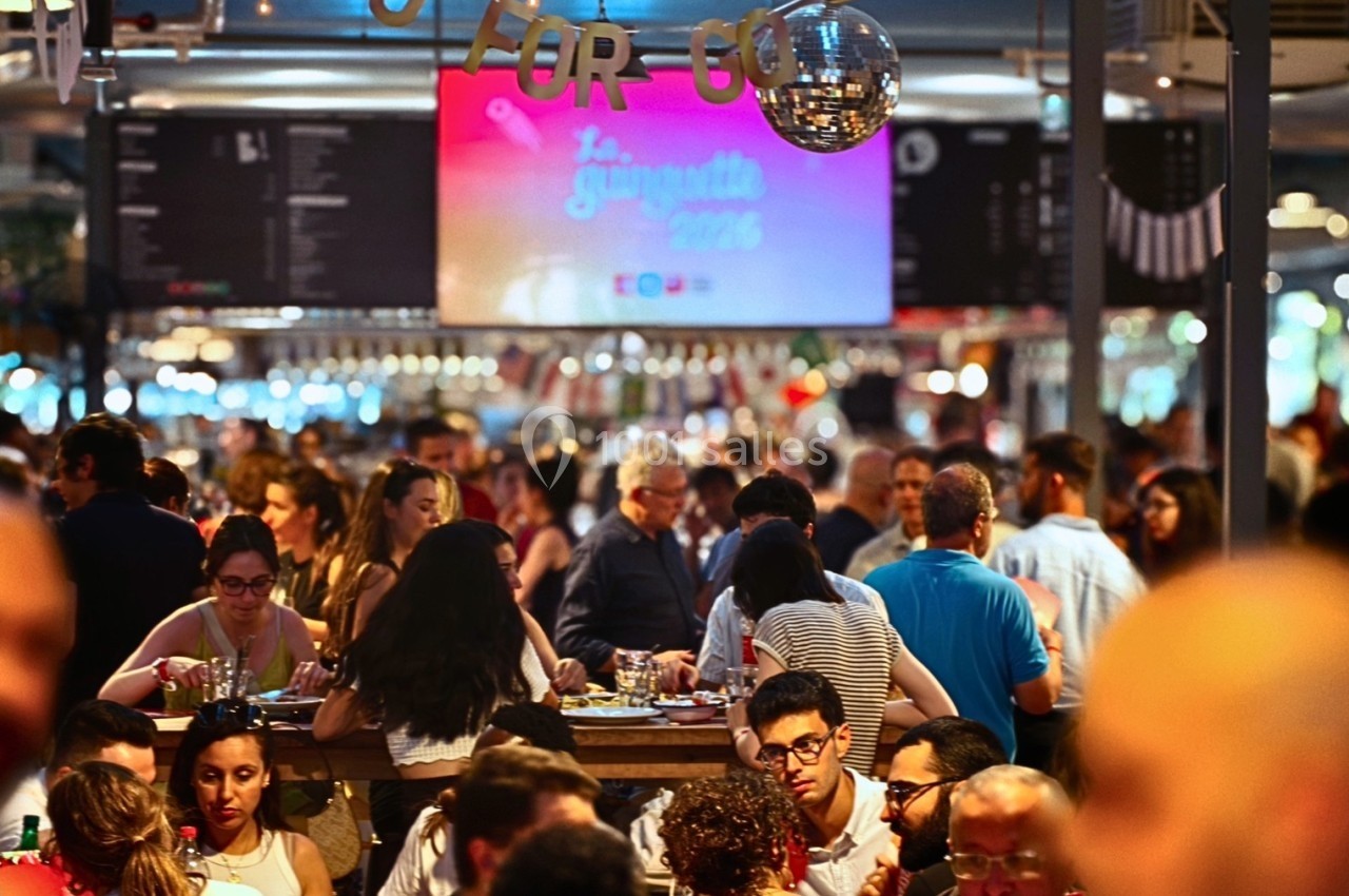 Salle bondée avec des personnes assises à des tables, éclairage chaleureux et écran affichant un événement en arrière-plan.