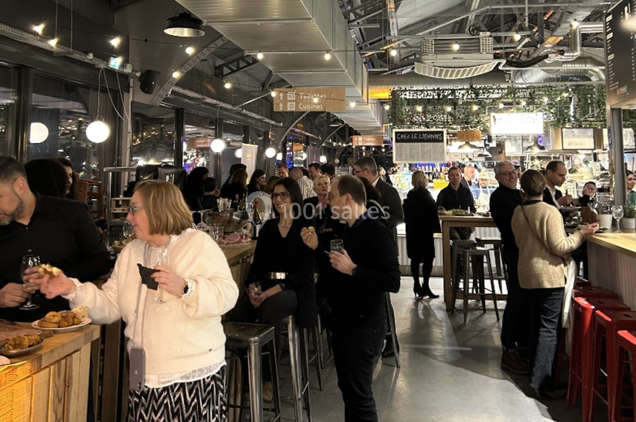 Des personnes discutent et dégustent des boissons dans un marché couvert animé, éclairé par des lumières chaleureuses.