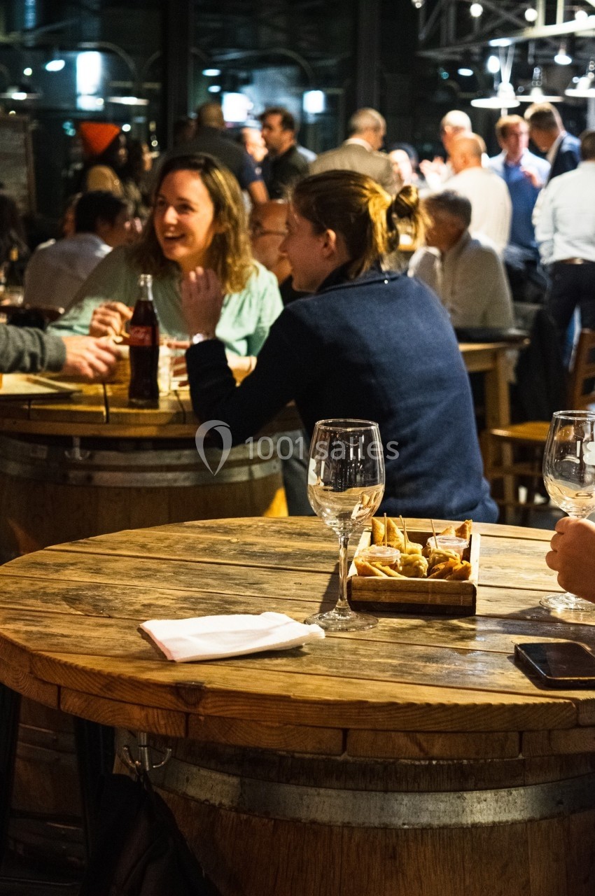 Des personnes discutent autour de tables en bois dans un bar animé, avec des verres et des amuse-bouches.