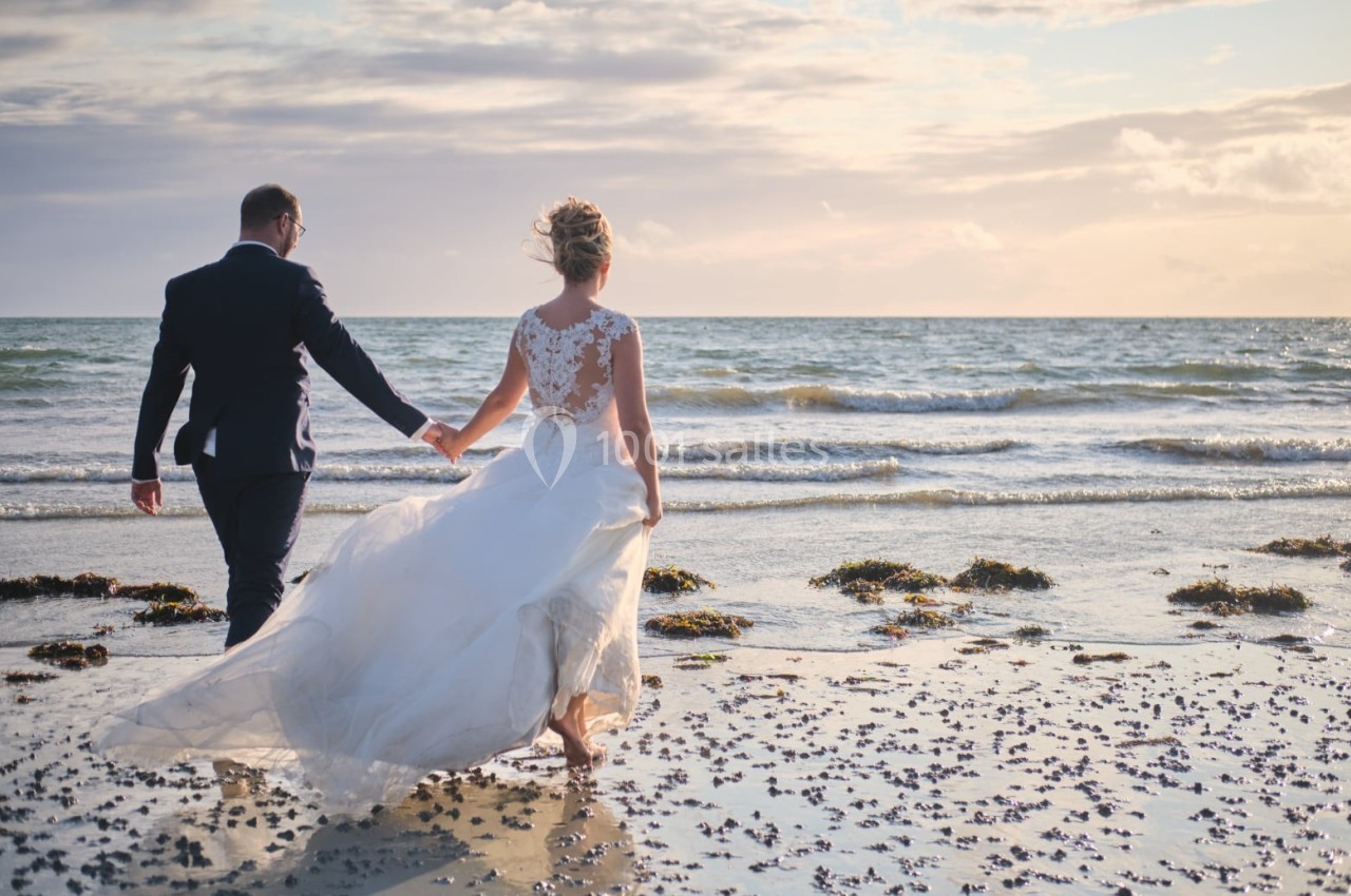 Un couple en tenue de mariage marche sur une plage au coucher du soleil, face à la mer.
