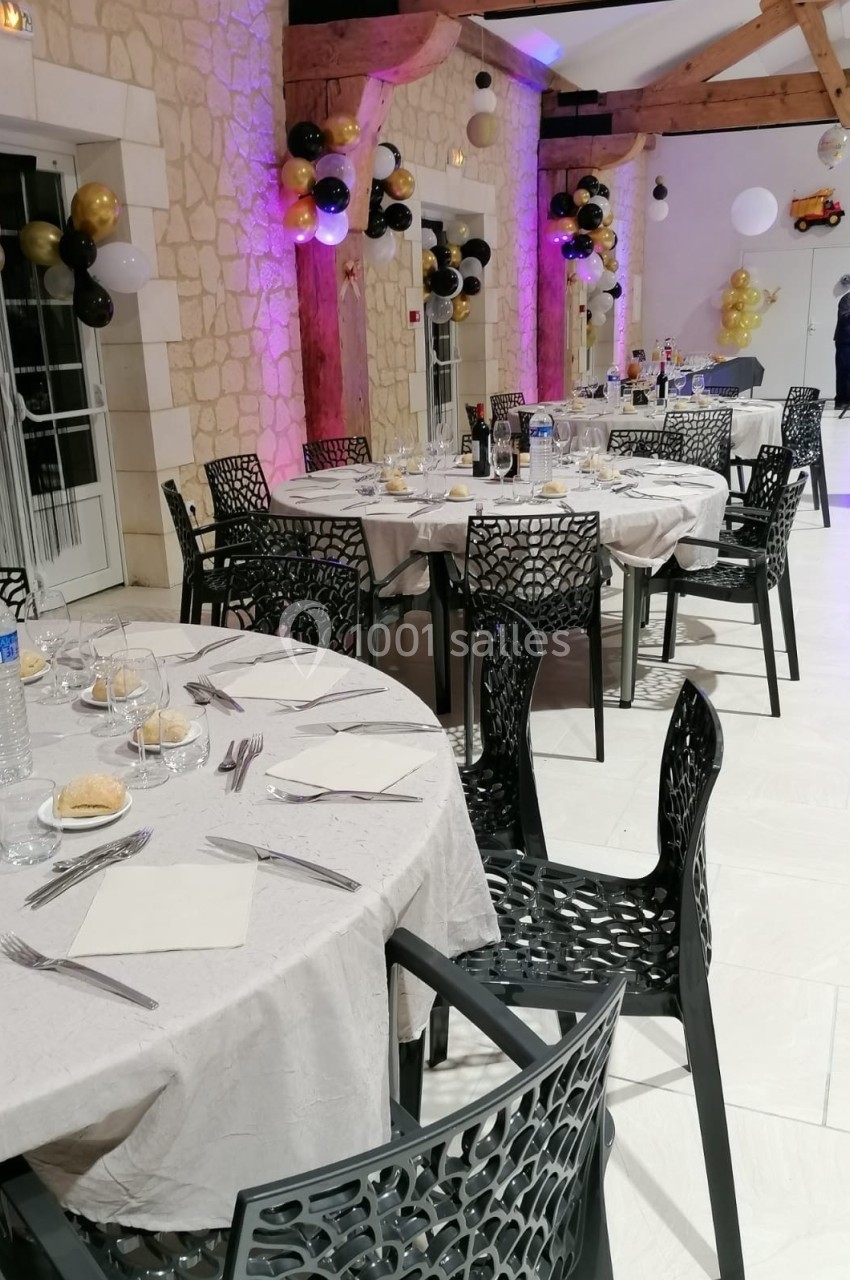 Salle décorée pour un événement avec tables dressées, chaises noires ajourées et ballons suspendus en arrière-plan.