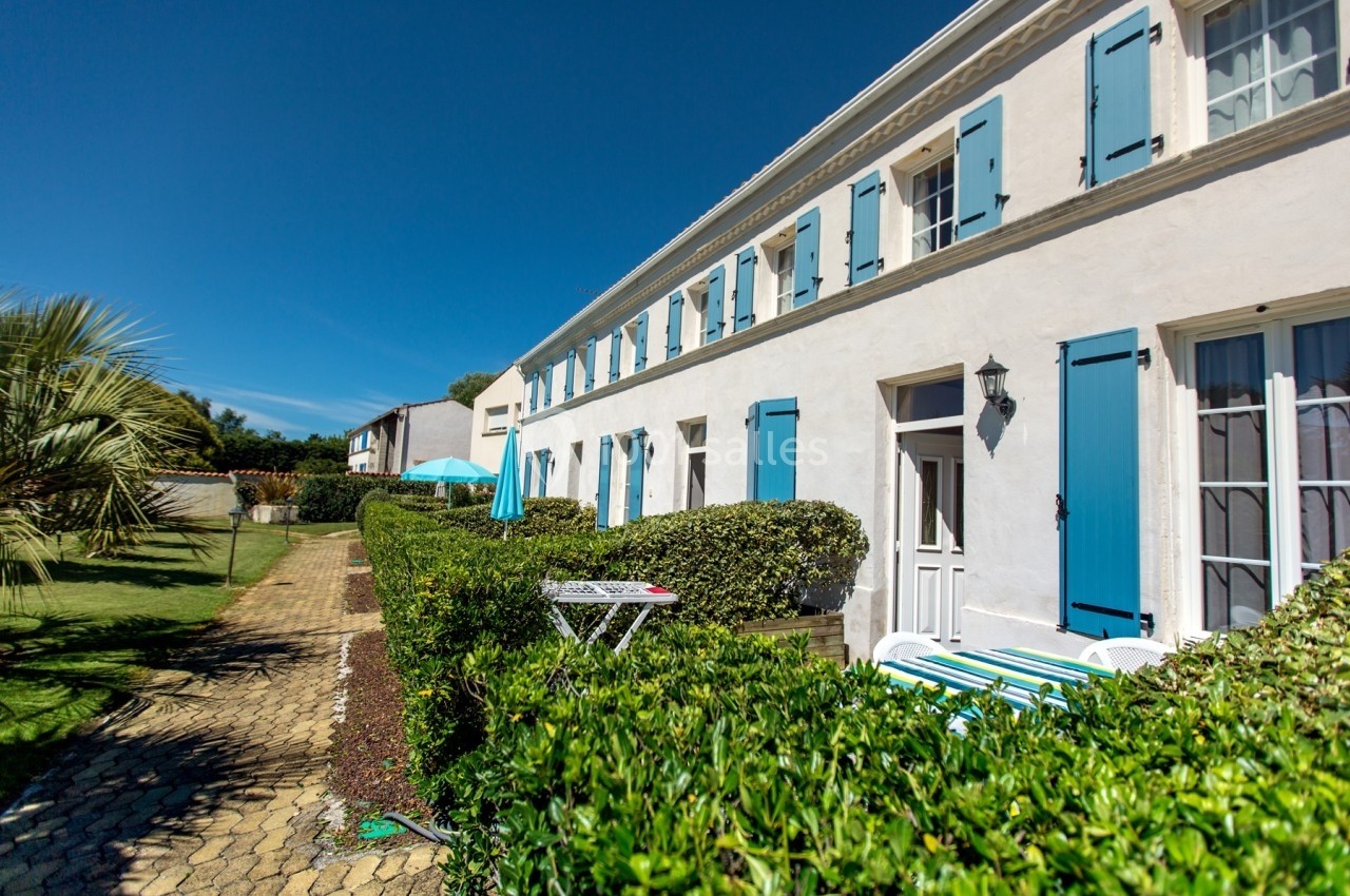 Façade d'une maison blanche avec volets bleus, entourée de haies et d'un jardin sous un ciel ensoleillé.