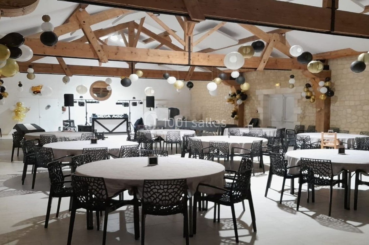 Salle de réception décorée avec des ballons noirs, blancs et dorés, tables rondes dressées et poutres apparentes.