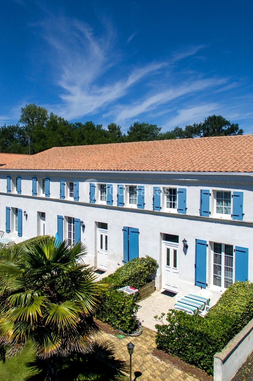 Grande maison blanche à volets bleus avec jardin arboré, sous un ciel bleu dégagé.