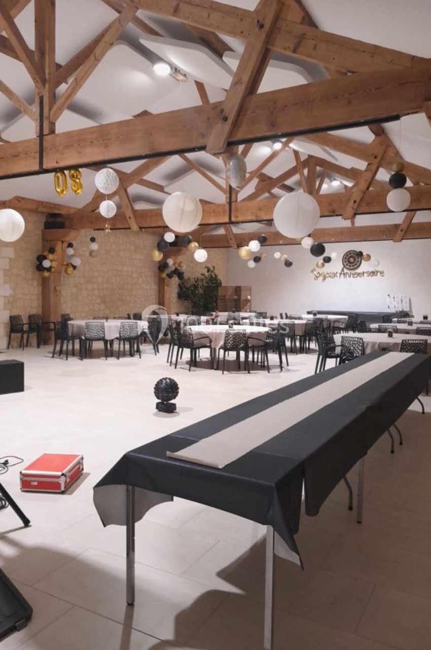 Salle de réception décorée pour un anniversaire, avec tables alignées, guirlandes et lanternes suspendues au plafond en bois.