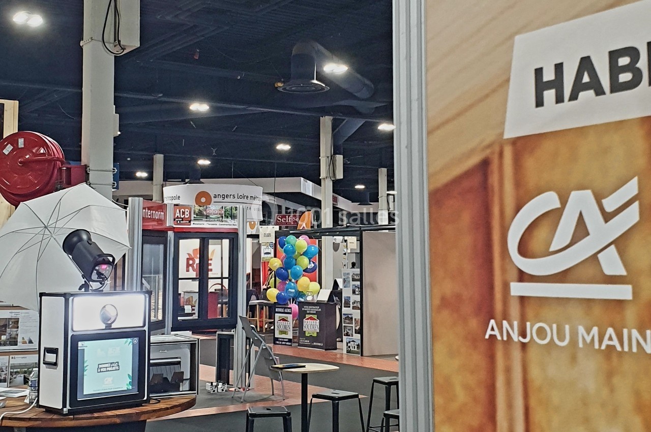 Borne photo avec éclairage et décor, installée dans un salon professionnel avec stands et visiteurs en arrière-plan.