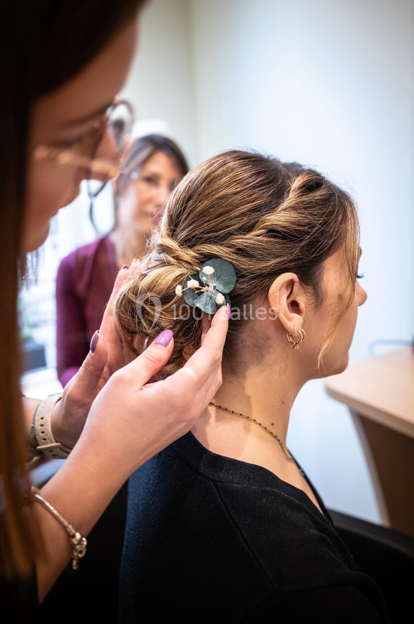 Une coiffeuse ajuste une barrette florale dans les cheveux attachés d'une femme, en intérieur.
