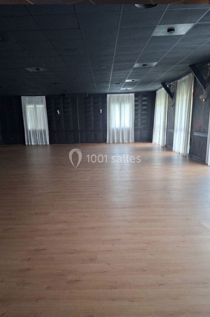 Salle vide avec parquet en bois clair, murs sombres et grandes fenêtres avec rideaux blancs laissant passer la lumière.