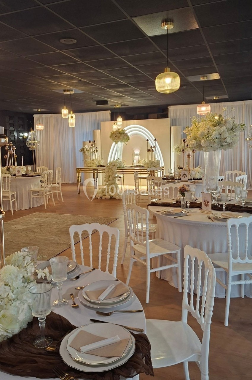 Salle de réception élégante décorée de fleurs blanches, tables rondes dressées et arche lumineuse en arrière-plan.