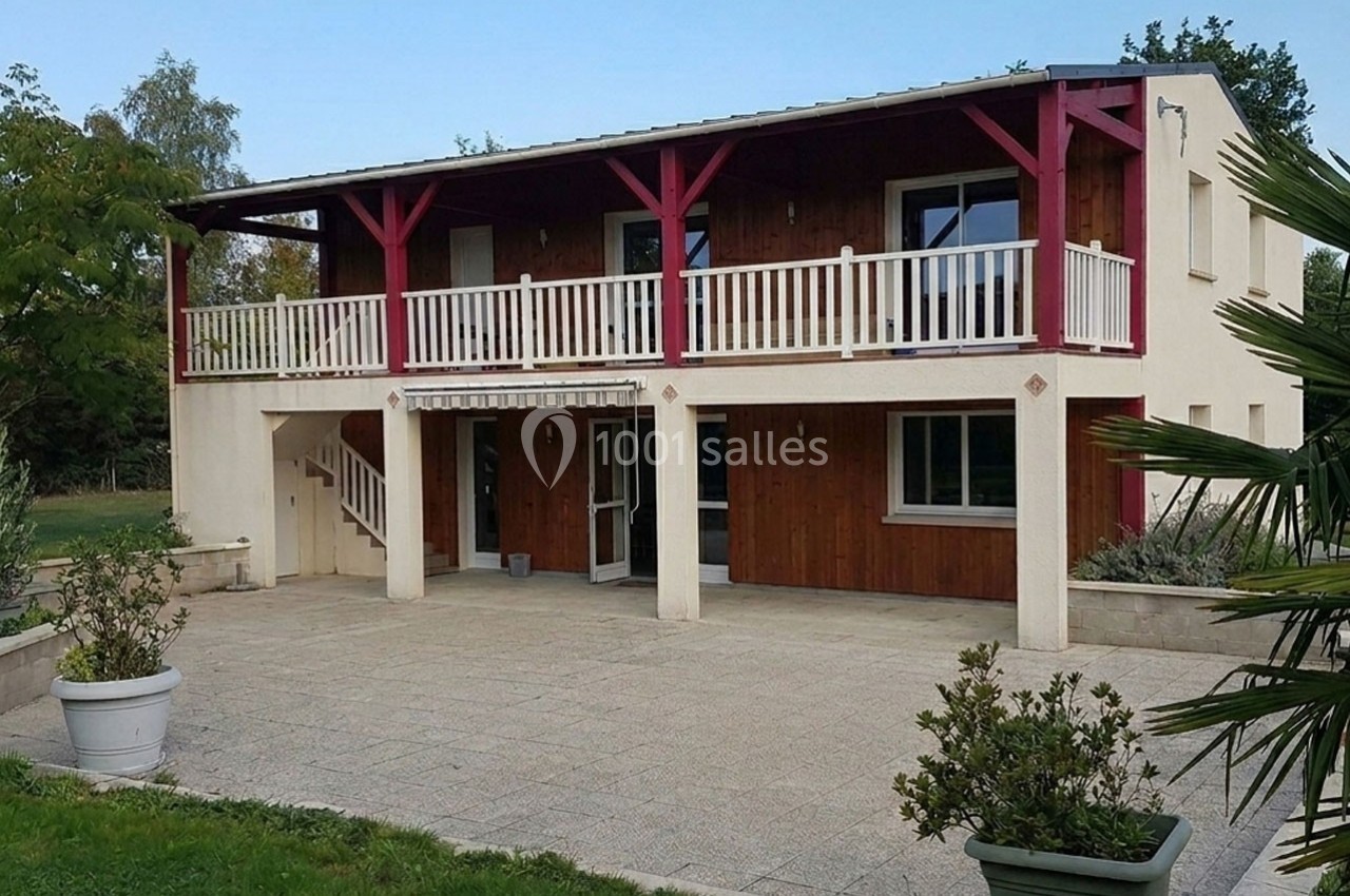 Maison à deux étages avec façade en bois, balcons et terrasse carrelée, entourée de végétation.