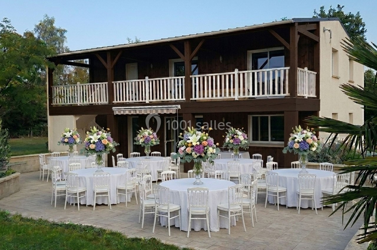 Terrasse aménagée avec des tables rondes dressées et décorées de grands bouquets de fleurs devant un bâtiment en bois.