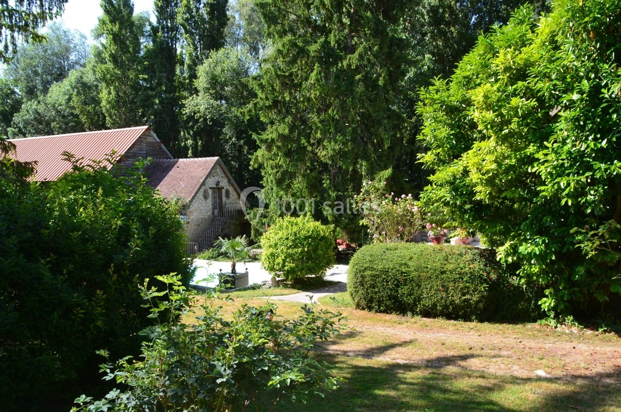 Jardin verdoyant avec pelouse, arbustes et arbres, maison en pierre avec toit en tuiles rouges en arrière-plan.