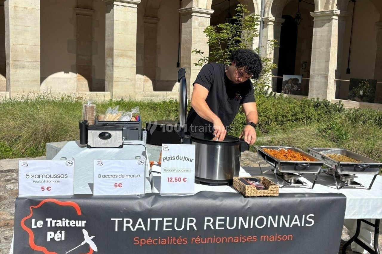 Un traiteur prépare des plats réunionnais derrière un stand avec des étiquettes de prix et des spécialités exposées.