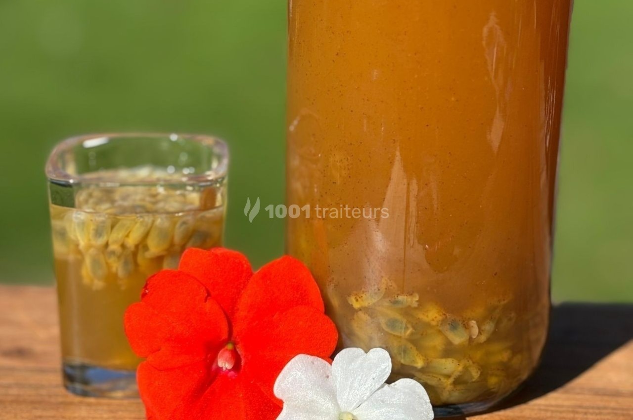 Boisson orange dans une carafe et un verre avec graines visibles, décorée de fleurs rouges et blanches sur une table en bois.