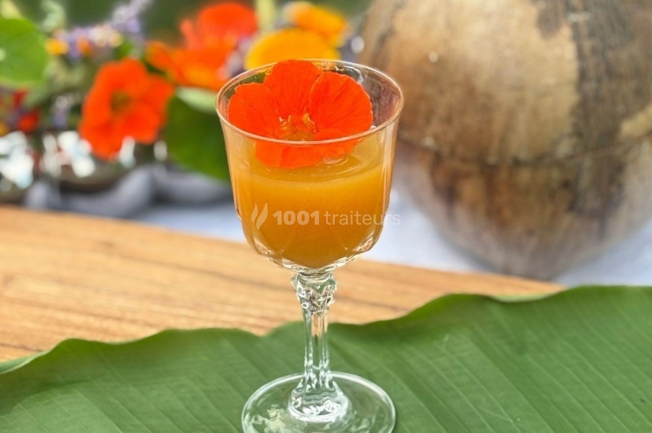 Verre de jus orange décoré d'une fleur comestible, posé sur une feuille de bananier avec un fond tropical.