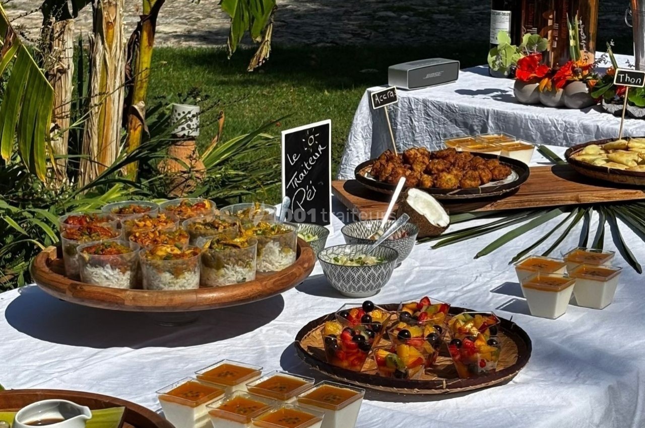Buffet en extérieur avec verrines, desserts, plats variés et décorations tropicales sur une nappe blanche.