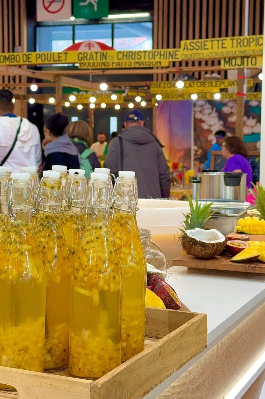 Bouteilles de jus tropical avec morceaux de fruits en avant-plan, stand de cuisine créole animé en arrière-plan.