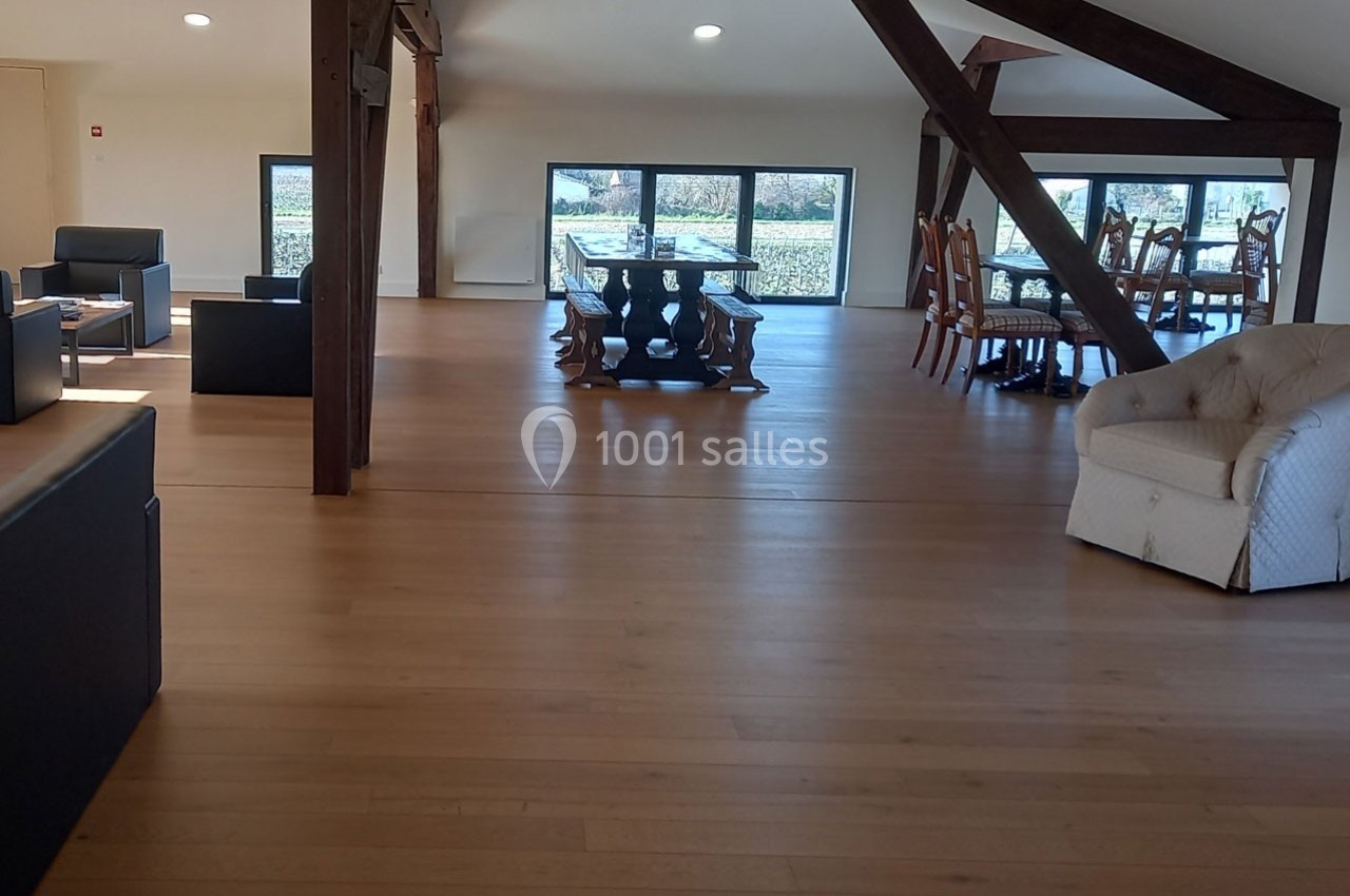 Salle spacieuse avec parquet en bois clair, poutres apparentes, canapés, et tables près de grandes fenêtres.