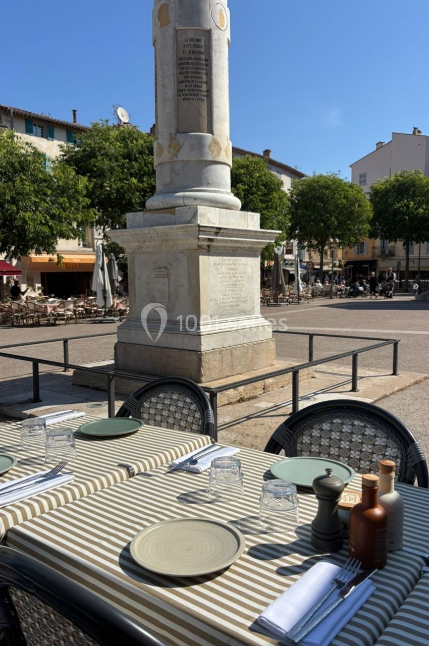 Tables dressées en terrasse avec vue sur une place ensoleillée et une colonne monumentale entourée d'arbres.