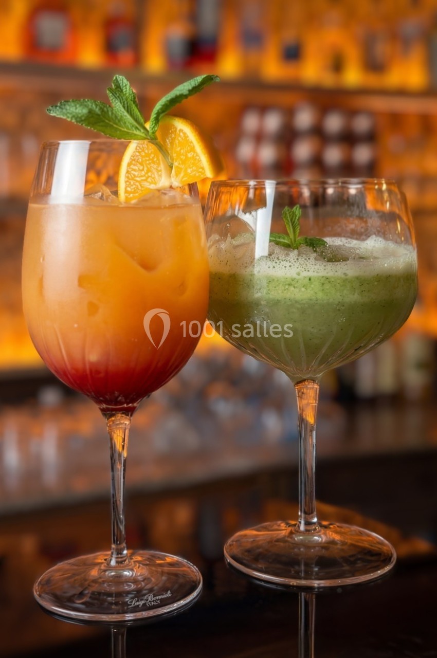 Deux cocktails colorés dans des verres à pied, l'un orange avec des tranches d'orange, l'autre vert avec une feuille de…