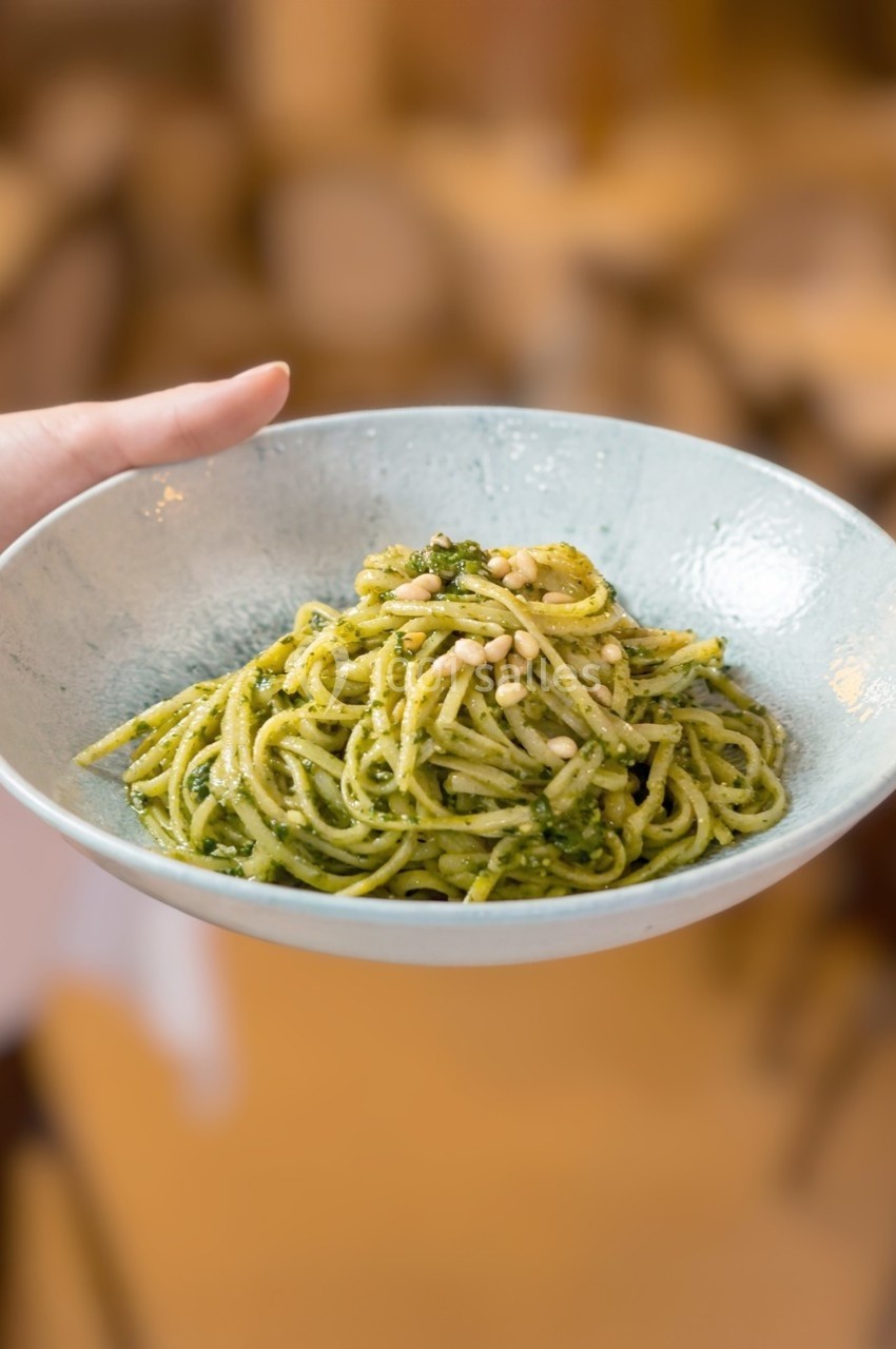 Assiette de spaghettis au pesto garnis de pignons de pin, tenue à la main dans un cadre flou de restaurant.