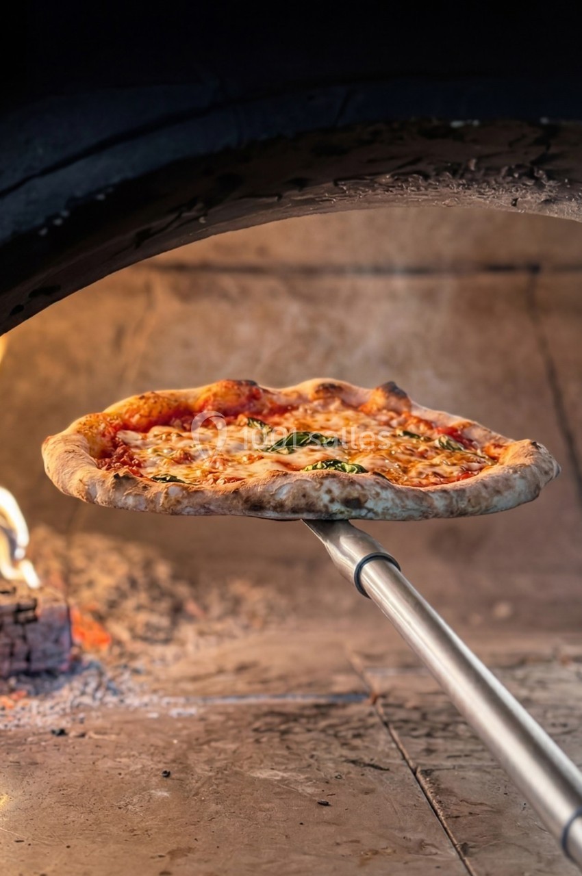 Pizza cuite au feu de bois sortie d'un four traditionnel sur une pelle en métal, avec des flammes en arrière-plan.