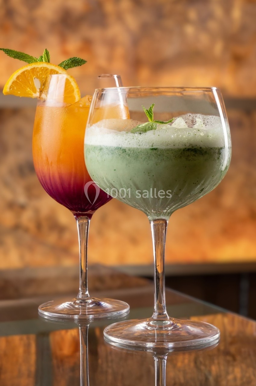 Deux cocktails colorés servis dans des verres à pied, l'un orange et rouge, l'autre vert, décorés de menthe et d'orange.