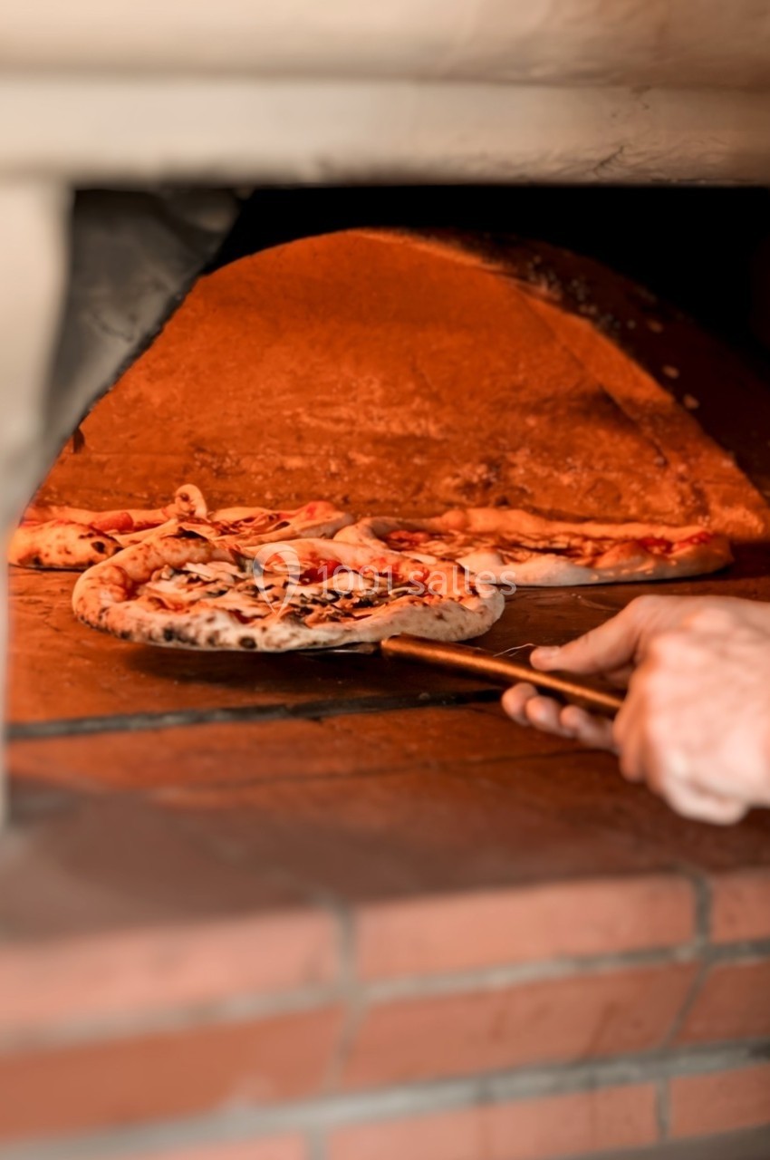 Pizzas en cours de cuisson dans un four à bois, une main utilise une pelle pour les manipuler.