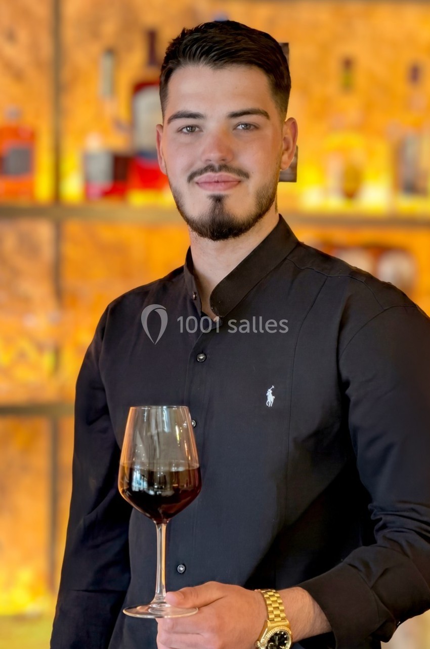 Un homme en chemise noire tient un verre de vin rouge devant un fond lumineux de bouteilles.