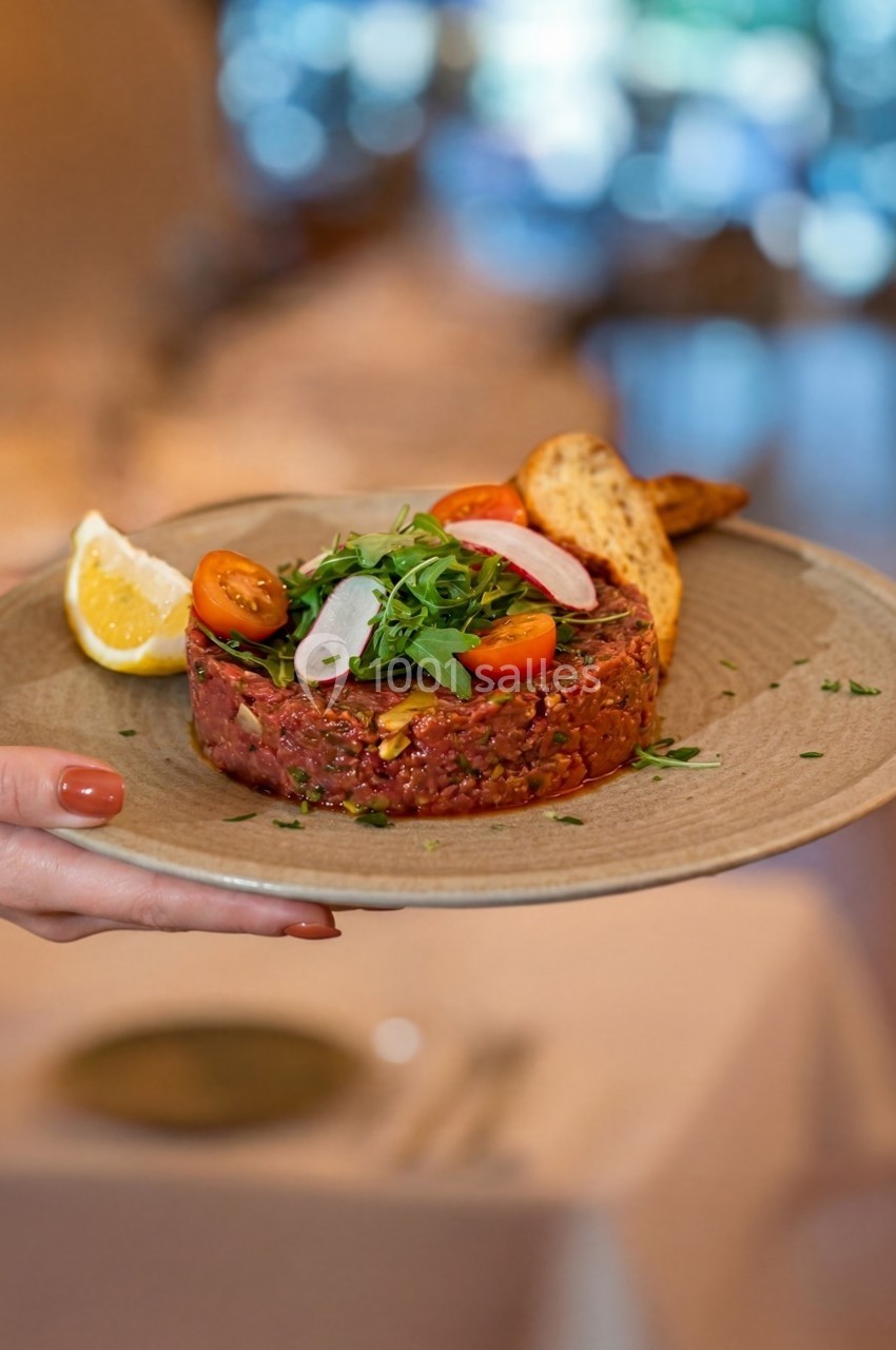 Assiette de tartare de viande garnie de roquette, tomates cerises, radis, citron et toasts, tenue par une main.