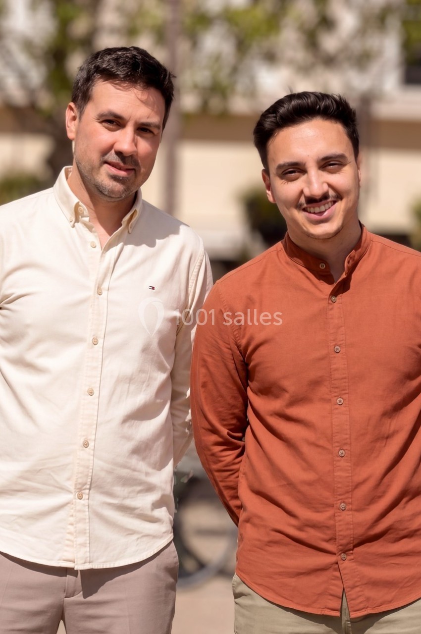 Deux hommes debout côte à côte, souriant, en extérieur avec un arrière-plan flou de bâtiments et de végétation.